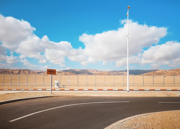 Strada vuota vicino al parcheggio dell'aeroporto di Eilat con segnale di uscita (traduzione in inglese e israeliano), montagne basse dietro la recinzione in lontananza