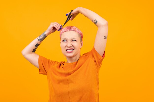 Straordinaria bella donna con i capelli corti rosa e grande tatuaggio sulla sua mano fare hairdress isolato su arancione