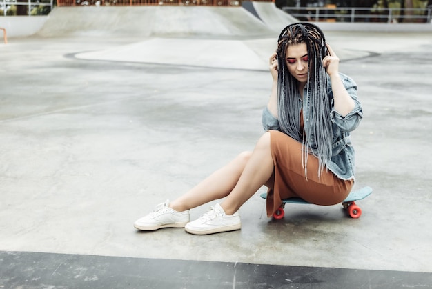 Straordinaria pattinatrice millenaria con trucco colorato Trecce africane si siede su un penny board e ascolta musica in cuffia allo skate park Concetto di stile di vita giovanile