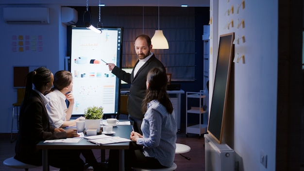 Strategia di marketing di brainstorming di un uomo d'affari maniaco del lavoro che lavora troppo nella sala riunioni dell'ufficio a tarda notte. Diversi uomini d'affari multietnici che guardano la presentazione finanziaria sul monitor