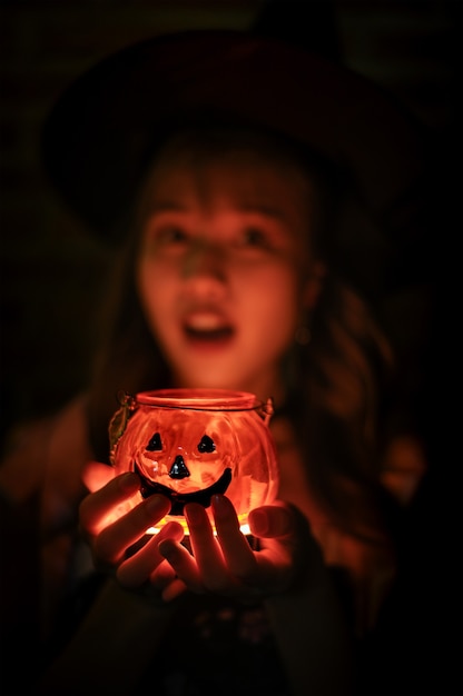 Strega affascinante che tiene la tazza della candela della zucca di Halloween alla luce rossa su fondo scuro