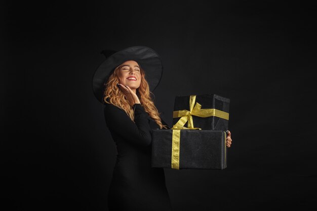 Strega di Halloween con un cappello nero su una parete nera che tiene i contenitori di regalo