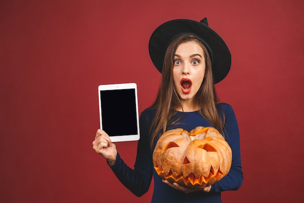 Strega di Halloween con uno schermo intagliato della compressa e della zucca - isolato su fondo rosso. Emotiva giovane donna in costume di Halloween.