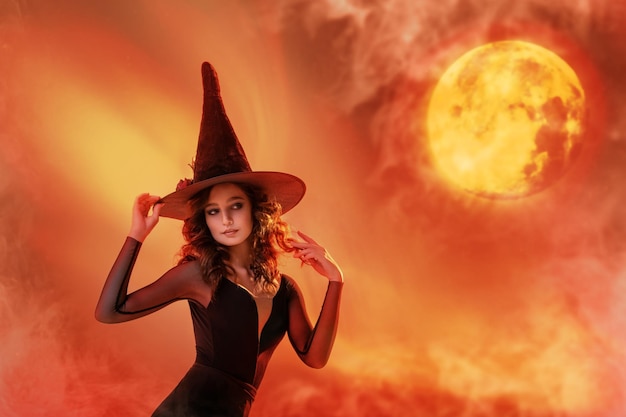 Strega di Halloween Una bella giovane donna con un cappello da strega evoca pratiche di stregoneria sullo sfondo di un tramonto Ampio design artistico di Halloween