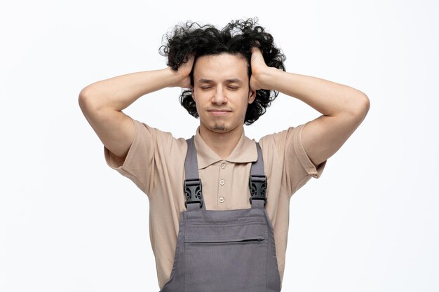 Stressato giovane operaio edile maschio che indossa l'uniforme tenendo le mani sulla testa con gli occhi chiusi isolati su sfondo bianco