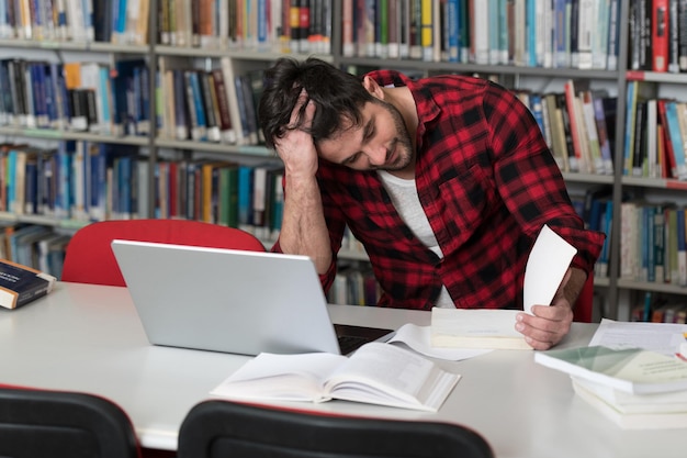 Stressato giovane studente maschio leggendo il libro di testo mentre era seduto in biblioteca