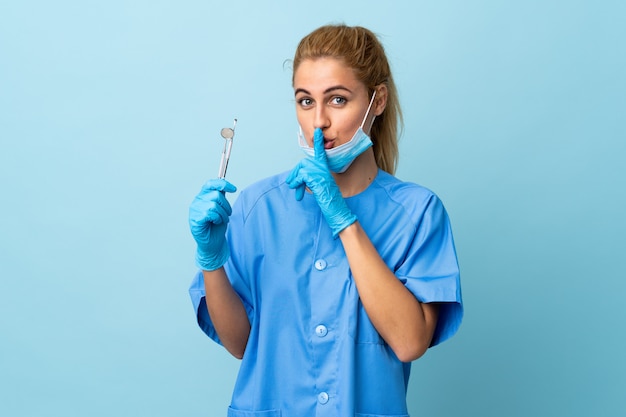 Strumenti di tenuta del dentista della giovane donna sopra la parete blu che fa gesto di silenzio