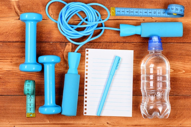 Strumenti per il fitness su fondo di legno marrone. Attrezzatura sportiva in colore blu ciano, vista dall'alto. Manubri, corda per saltare, centimetro, penna, quaderno e bottiglia per dimagrire. Concetto di palestra e stile di vita sano.