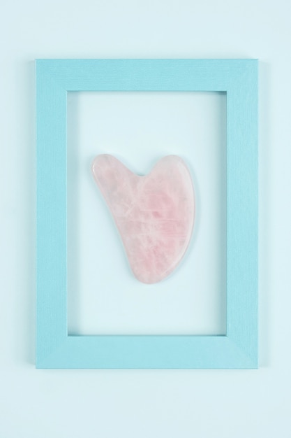 Strumento di massaggio giada Gua sha da quarzo rosa cristallo in cornice su sfondo blu. Massaggio viso antietà per un trattamento lifting e tonificante naturale a casa. Concetto di bellezza Vista dall'alto.