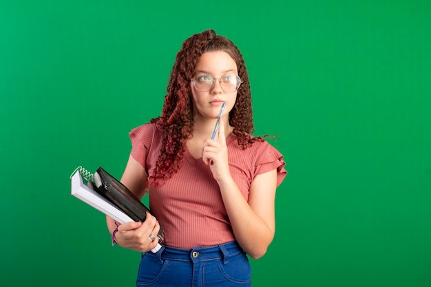 Studente adolescente con gli occhiali che indossa una borsa da scuola e porta quaderni in posa in una foto in studio con uno sfondo verde ideale per il ritaglio