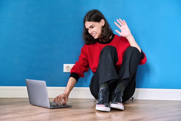 Studente adolescente in cuffia che utilizza un laptop per una videochiamata in conferenza seduto sul pavimento su sfondo blu Concetto di tecnologia giovanile per l'apprendimento online di Eeducation