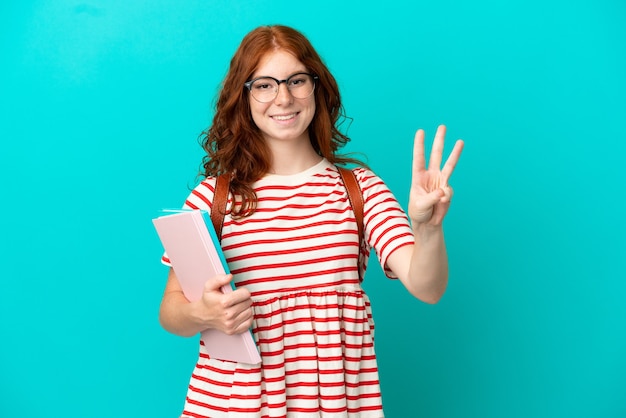 Studente adolescente ragazza rossa isolata su sfondo blu felice e contando tre con le dita