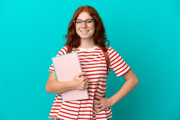 Studente adolescente ragazza rossa isolata su sfondo blu in posa con le braccia all'anca e sorridente