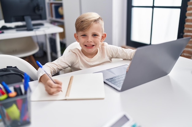 Studente adorabile del bambino che utilizza la scrittura del computer portatile sul taccuino all'aula