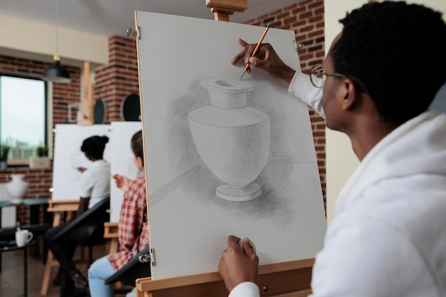 Studente afroamericano che frequenta una lezione d'arte lavorando a un'opera d'arte creativa che disegna un modello di vaso utilizzando la matita grafica nello studio della creatività. Disegno di schizzo della squadra multietnica. Concetto creativo