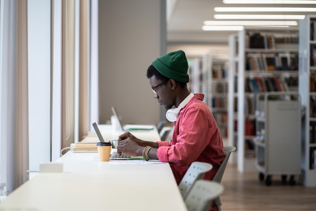 Studente afroamericano concentrato seduto a una scrivania con un laptop che usa il database della biblioteca online