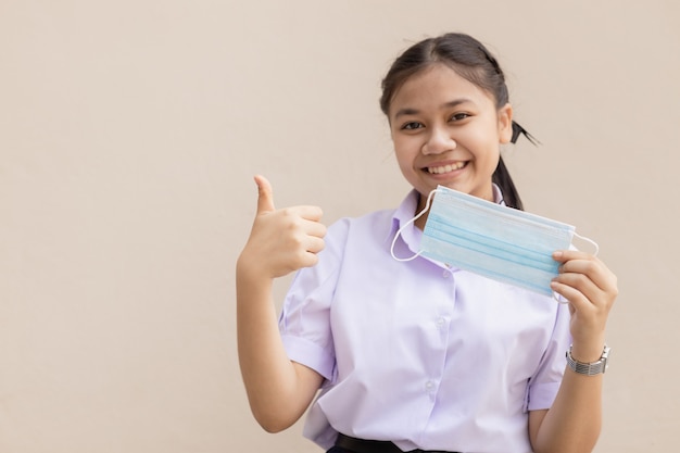 Studente asiatico carino in uniforme felice con la maschera facciale pollice in alto per una buona salute.