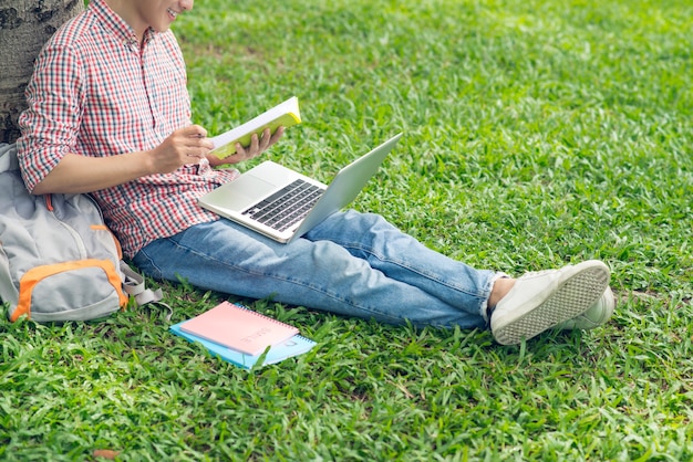 Studente asiatico che legge un libro mentre è seduto sull'erba verde