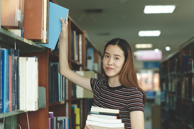 Studente asiatico che sceglie libro nello scaffale in biblioteca.