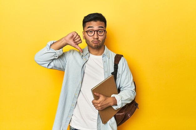 Studente asiatico con zaino e notebook sullo sfondo giallo dello studio che mostra i pollici di un gesto di antipatia