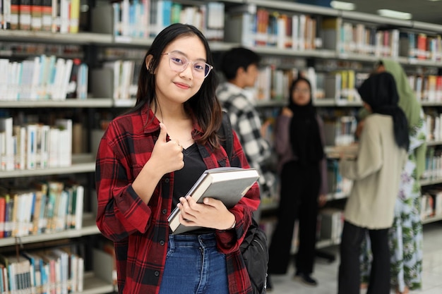 Studente asiatico in piedi con sicurezza e dando il pollice in su mentre sorride e tiene il libro in biblioteca