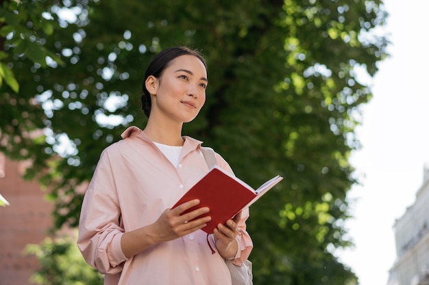 Studente asiatico pensieroso che studia il libro di lettura che distoglie lo sguardo all'aperto