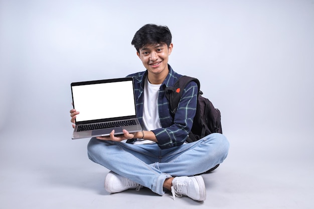 Studente asiatico seduto con le gambe incrociate sul pavimento e che mostra lo schermo bianco del portatile per il mockup