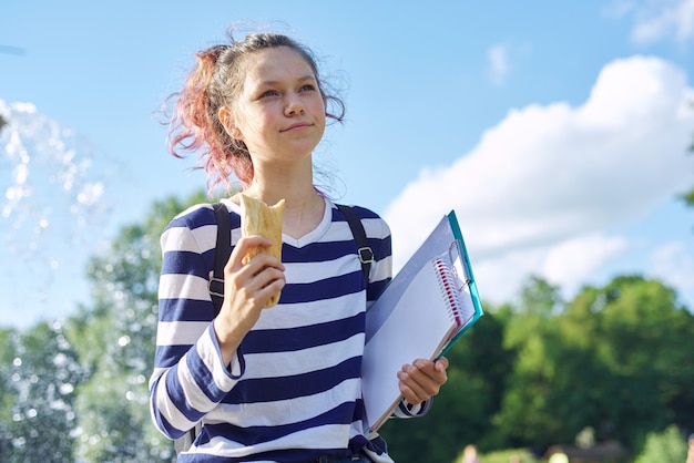 Studente che cammina all'aperto con zaino, quaderni e mangia panino