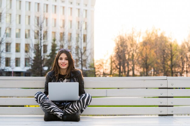 Studente che si siede sul banco che ascolta la musica e che per mezzo del computer portatile contro il campus universitario