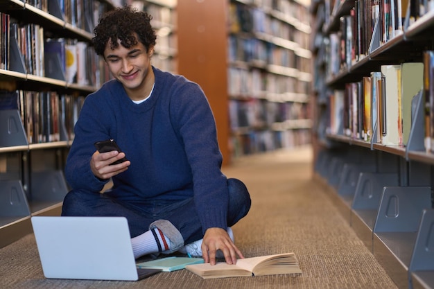 Studente che trascorre del tempo in biblioteca utilizzando il telefono cellulare e il laptop alla ricerca di informazioni per il progetto di diploma