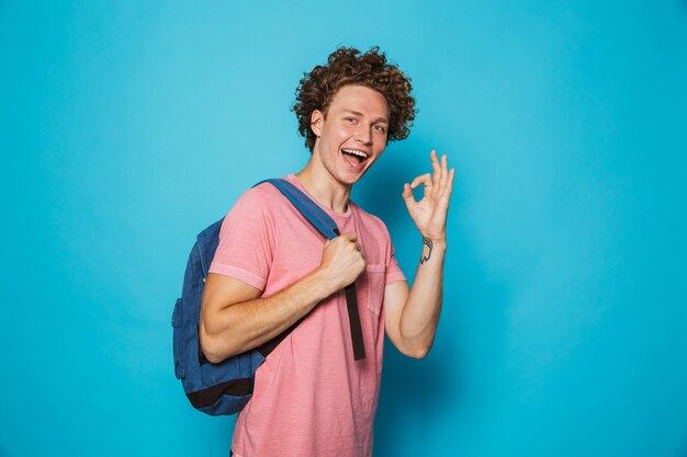 Studente con capelli ricci che indossa abbigliamento casual e zaino che sorride e che mostra segno giusto