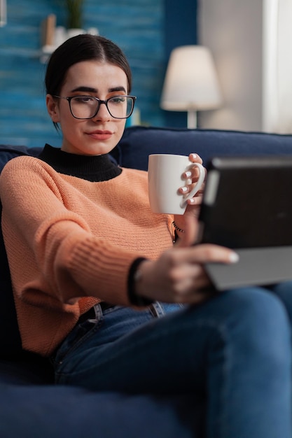 Studente con gli occhiali sul divano che tiene la tazza guardando il display del tablet. Influencer sul divano in videochiamata mentre beve caffè o tè. Creatore di contenuti in streaming video su dispositivo digitale.