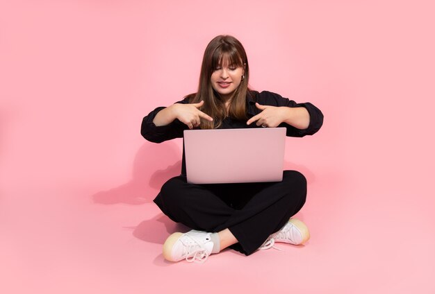 Studente dai capelli castani Ragazza in abiti casual che tiene il computer portatile seduto su sfondo rosa. Concetto di formazione online. Colpo Dello Studio. Guardando la fotocamera. mostrando al laptop