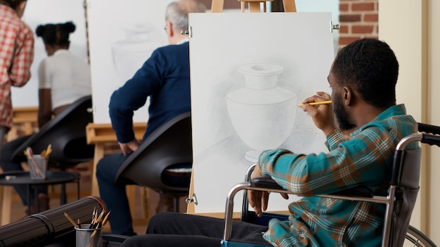 Studente di classe d'arte in sedia a rotelle che usa la matita su tela per disegnare un modello di vaso per la crescita delle abilità personali. Impara la pratica del disegno per creare disegni e schizzi d'arte nella classe della scuola artistica.