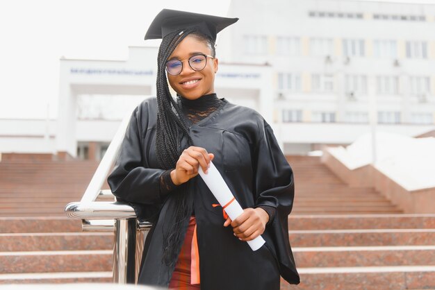 Studente di college abbastanza africano in cappello di laurea e abito davanti all'edificio scolastico