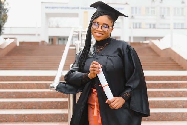 Studente di college abbastanza africano in cappello di laurea e abito davanti all'edificio scolastico