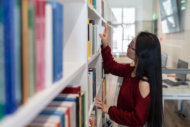 Studente di college asiatico in biblioteca concentrata ragazza che raccoglie libri per prepararsi per l'esame all'università