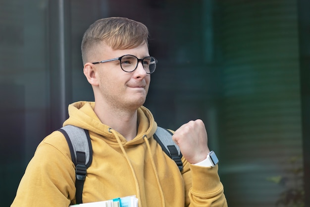 Studente di college bello successo positivo felice, allievo allegro, giovane che sorride con i libri, libri di testo, occhiali guarda l'elenco dei risultati. Il ragazzo intelligente ha superato l'esame universitario, celebrando la vittoria, il trionfo