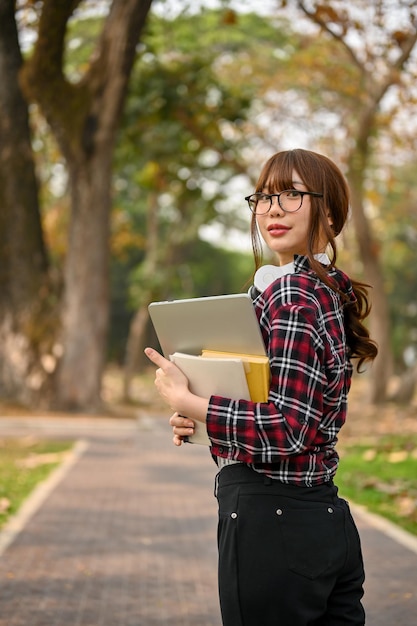 Studente di college femminile asiatico che tiene il suo computer portatile e libro che camminano lungo il parco