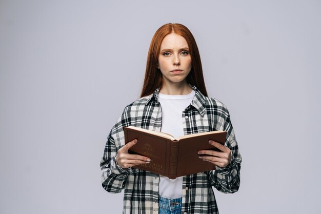 Studente di college giovane donna seria in possesso di libri aperti e guardando la fotocamera su sfondo grigio isolato Modello di signora piuttosto rossa che indossa abiti casual che mostra emotivamente le espressioni facciali