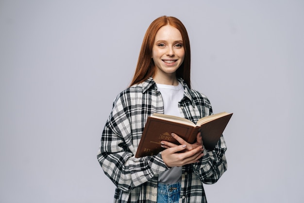 Studente di college sorridente attraente della giovane donna che tiene libro e che esamina macchina fotografica