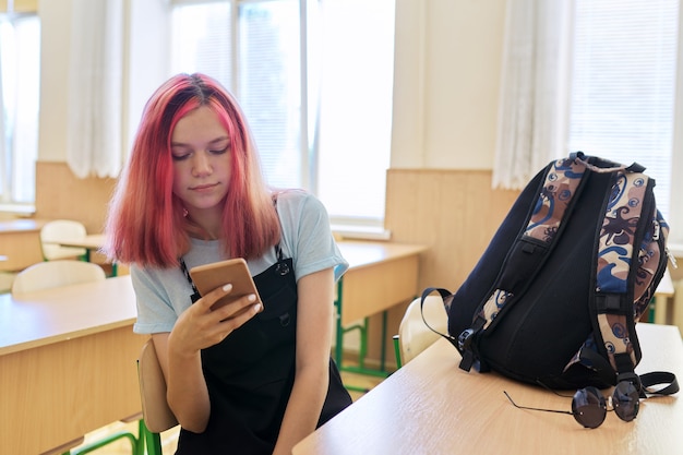 Studente di scuola adolescente ragazza alla moda, in classe con lo smartphone in mano, seduto alla scrivania in pausa tra le lezioni