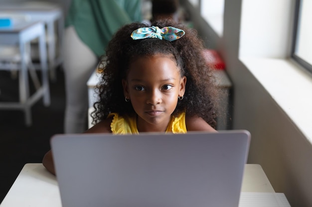Studente elementare afroamericana che guarda via mentre è seduta con il portatile durante la lezione di computer