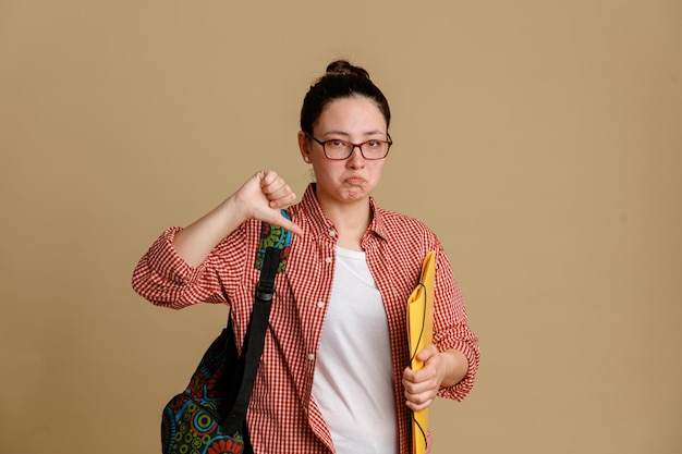 Studente giovane donna in abiti casual con gli occhiali con lo zaino che tiene la cartella guardando la fotocamera dispiaciuto e offeso che mostra il pollice in giù in piedi su sfondo marrone