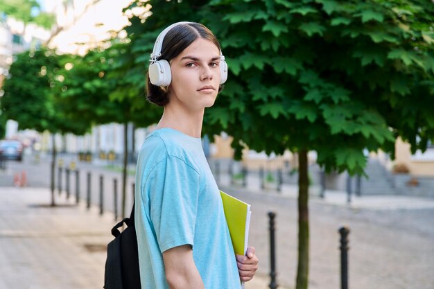 Studente maschio adolescente sorridente con libri di testo che guarda l'obbiettivo all'aperto Bel ragazzo 17 18 anni con le cuffie dello zaino sulla strada della città Concetto di stile di vita della gente di educazione giovanile