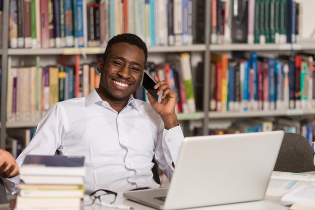 Studente maschio africano che parla al telefono nella profondità del campo poco profonda della biblioteca