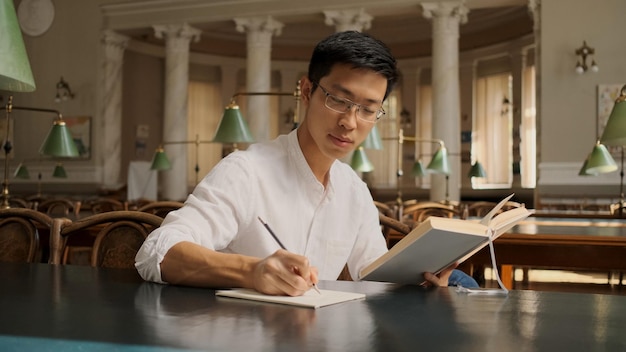 Studente maschio asiatico attraente con un libro di testo che prende appunti di studio in biblioteca Giovane ragazzo sicuro di sé che studia all'università