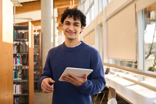 Studente maschio ispanico bello con la compressa che sorride alla macchina fotografica che visita un campus moderno della biblioteca