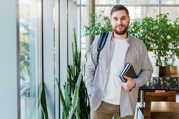 Studente nel riaprire il corridoio del campus universitario vicino alla finestra. Adolescente caucasico con i libri di testo dei taccuini dello zaino. Uomo barbuto bello sorridente. Libero professionista in un moderno ufficio di coworking con piante.