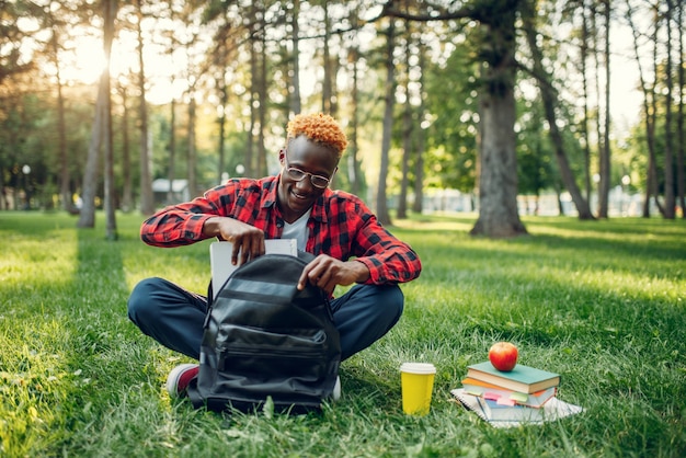 Studente nero con zaino seduto sull'erba nel parco estivo.